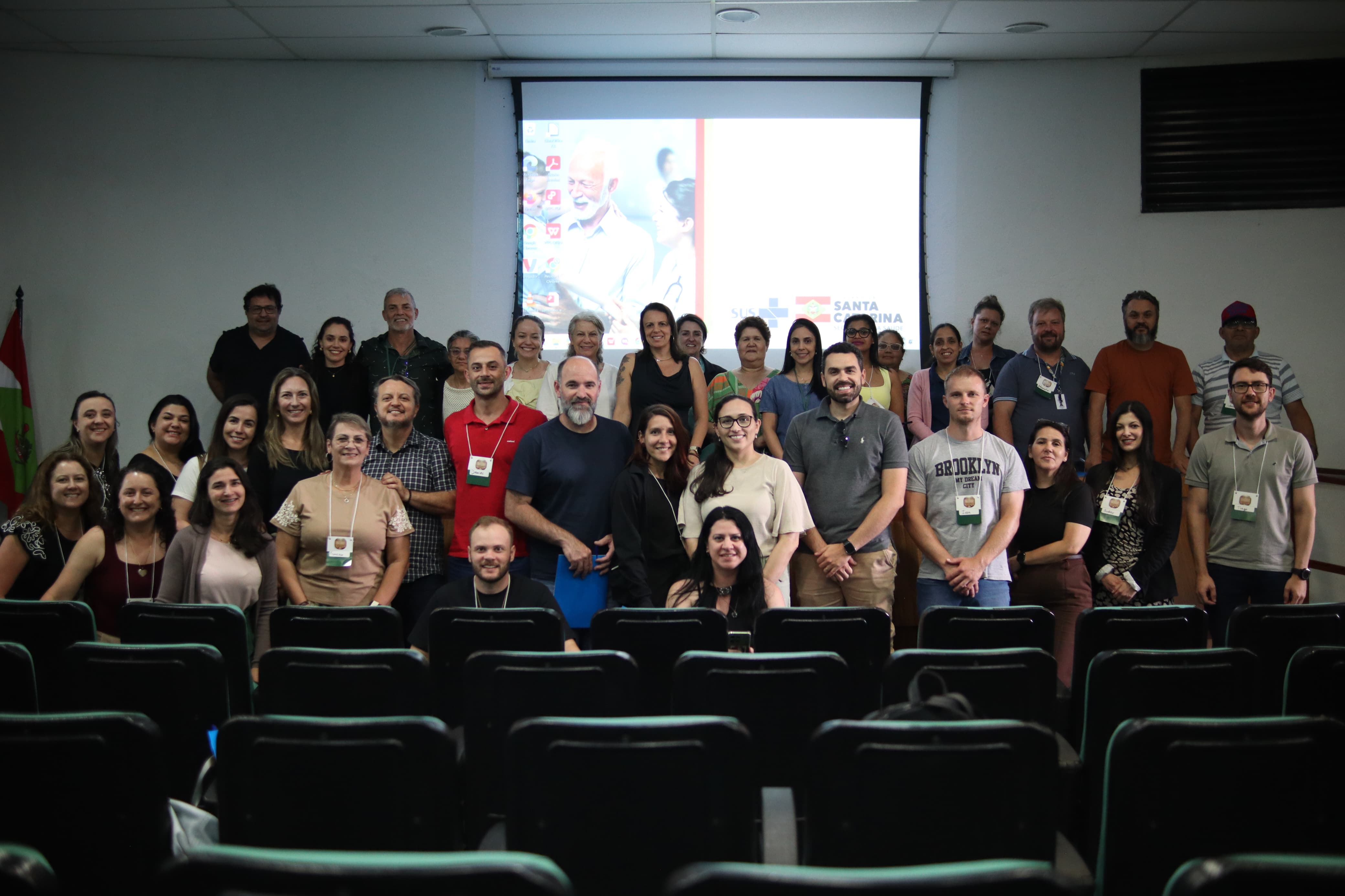 Grupo de participantes reunido em um auditório, posando para foto durante atividade ou capacitação. Ao fundo, há uma tela de projeção com apresentação institucional da Secretaria de Estado da Saúde de Santa Catarina, e à frente aparecem as cadeiras do auditório.