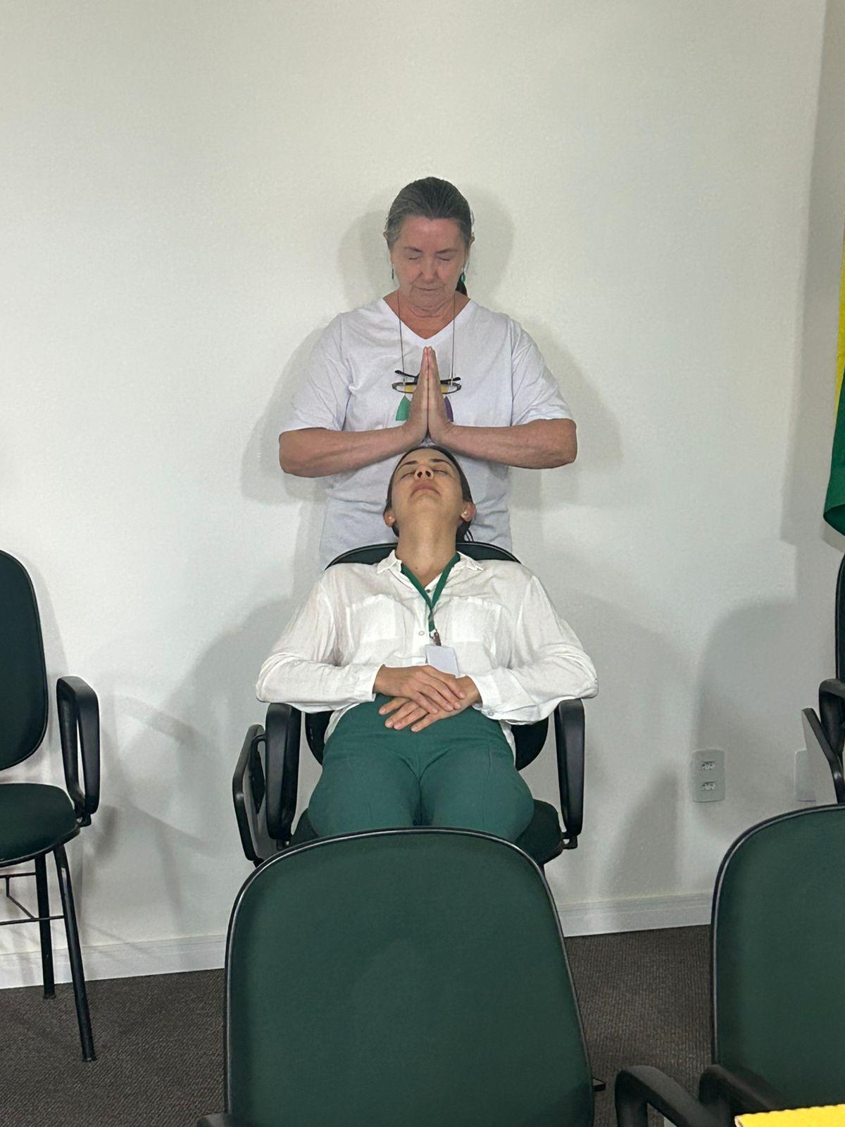 Profissional realiza atividade de relaxamento e cuidado terapêutico com servidora sentada em cadeira reclinada, em ambiente interno. A participante está com os olhos fechados enquanto a facilitadora posiciona as mãos sobre sua cabeça, em momento de acolhimento e bem-estar durante ação da campanha Abril Verde.