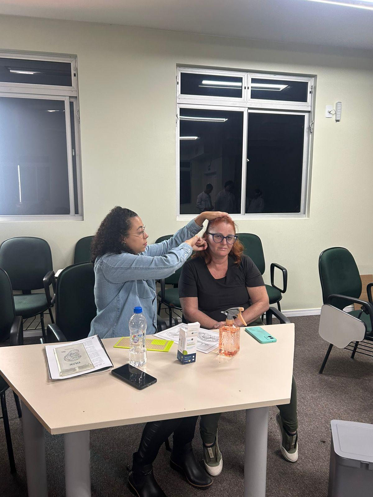 Profissional aplica técnica de auriculoterapia em participante sentada durante atividade de promoção da saúde em ambiente interno. Sobre a mesa há materiais utilizados na prática integrativa, como cartilhas, sementes terapêuticas e itens de apoio ao atendimento.