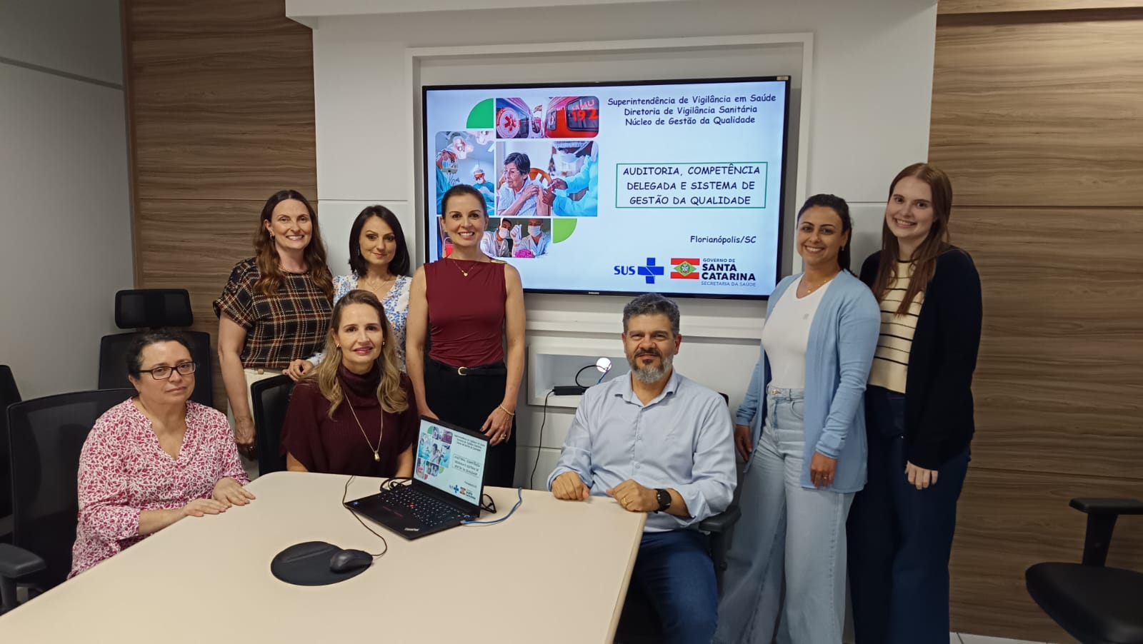 Equipe da Vigilância Sanitária reunida em sala de reunião, com sete mulheres e um homem, posando ao redor de uma mesa com notebook. Ao fundo, tela exibe apresentação com o tema auditoria, competência delegada e sistema de gestão da qualidade, com identificação da Superintendência de Vigilância em Saúde e Governo de Santa Catarina.