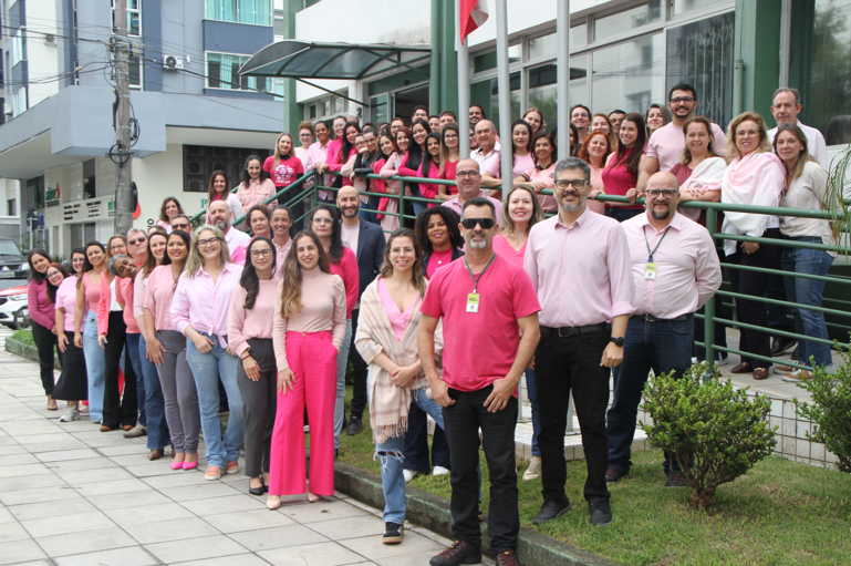 Servidores e servidoras da Secretaria de Estado da Saúde de Santa Catarina reunidos em frente ao prédio da instituição, vestindo diferentes tons de rosa em apoio à campanha Outubro Rosa. O grupo posa sorridente nas escadas e na calçada, com bandeiras hasteadas e edifícios ao fundo.