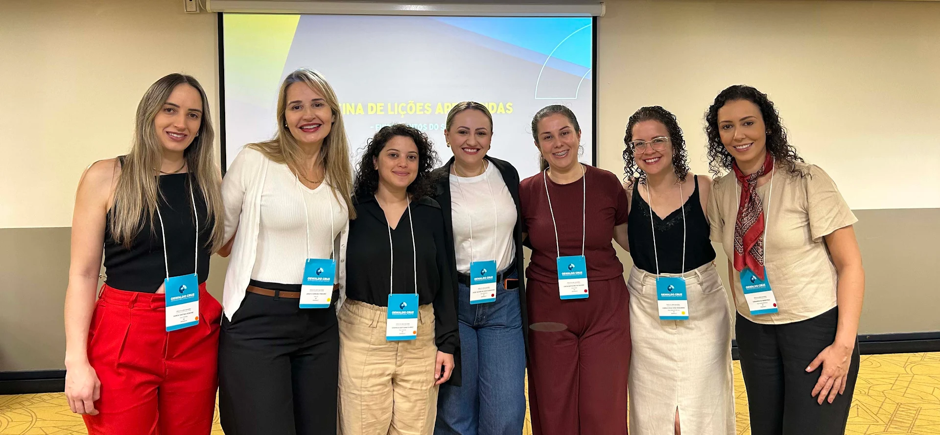 Grupo de sete mulheres em pé, alinhadas lado a lado, sorrindo para a câmera em um ambiente de evento ou conferência. Todas usam crachás azuis pendurados no pescoço. Ao fundo, há uma tela de projeção com parte de um título visível: “OFICINA DE LIÇÕES APRENDIDAS”. O local tem paredes claras e piso amarelo com padrão geométrico.